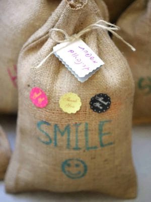 EFI_2317-scaled-scaled שקית (פאו'צ) מיוטה gift bag קשורה בחוט, מעוטרת במדבקות צבעוניות ותגית, עם הכיתוב SMILE ופרצוף סמיילי כחול בחזית.